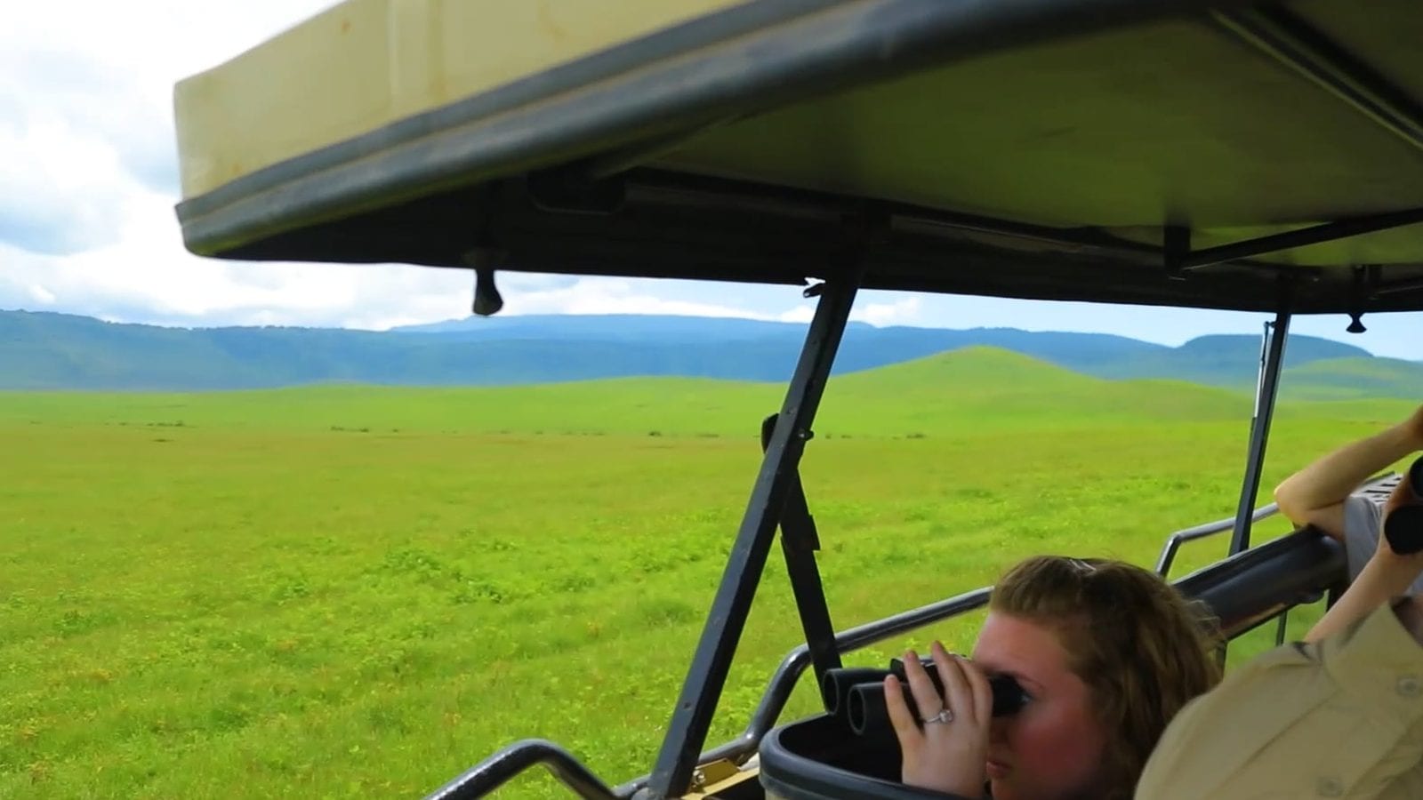 Gäste mit Ferngläsern unter dem aufgestellten Dach eines Safari-Fahrzeugs vor grüner Ebene